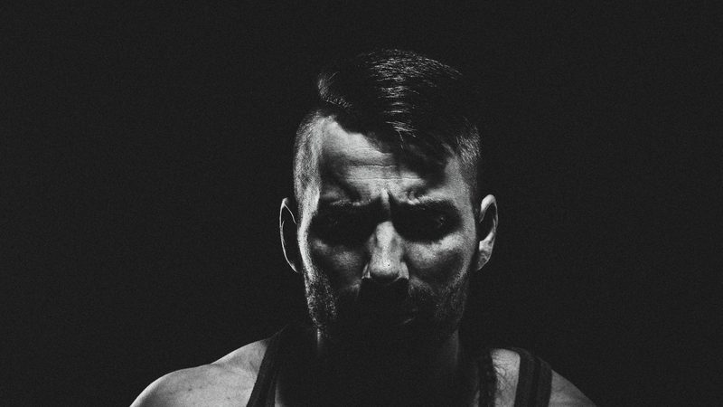 Bearded athlete in a black tank top lit from one side against black