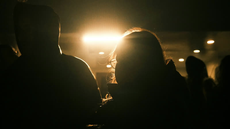 Two silhouettes in a dark venue, backlit by a single warm light.