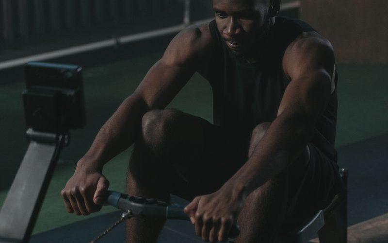 Athlete mid-stroke on a rowing machine in a dim gym, light from the side