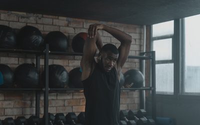 Athlete stretching an arm overhead in a dim gym beside a rack of medicine balls
